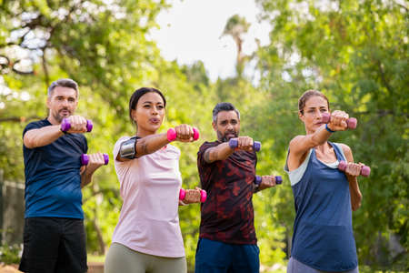Group of mature friends exercise using dumbbells at park. Team of four middle aged people lifting weights outdoor. Determined men and fit women using dumbbell for workout in park together.の写真素材