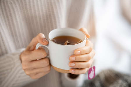 Woman wearing sweater and holding a warm cup of freshly brewed tea. Close up of woman hands holding mug of hot detox infusion in morning. Girl with cozy cloths relaxing at home during tea time in cold winter day.の写真素材