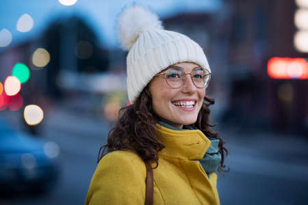 Smiling young woman wearing warm hat with eyeglasses on city street waiting for the bus. Happy girl wearing yellow coat and standing outdoors on winter evening at bus stop. Carefree happy girl in cold urban center during dusk.の写真素材