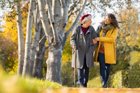 Granddaughter walking with senior woman in park wearing winter clothing. Old grandmother with walking cane walking with lovely caregiver girl in sunny day. Happy woman and smiling grandma walking in autumn park.の写真素材