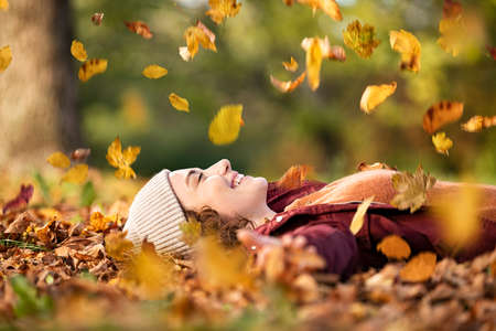 Cheerful young woman lying over leaves with closed eyes and smiling. Woman lying over autumn ground relaxing at park. Casual joyful girl having fun throwing leaves in autumn park with copy space.の写真素材