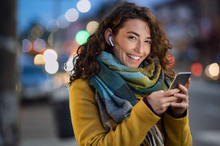 Young beautiful woman using her mobile phone at dusk in the city while listening music through earphones. Smiling girl sending message from cellphone in urban environment. Happy woman using smartphone to do a video call on a winter day while looking at camera.の写真素材