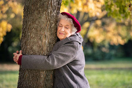 Beautiful senior woman hugging tree in the park. Mature woman hugging a tree and smiling outdoor. Senior lady with grey hair wearing red cap and grey coat embracing a trunk with eyes closed during autumn.の写真素材
