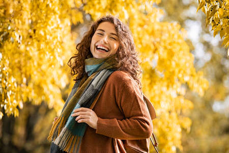 Carefree young woman wearing winter scarf with a big grin on face at park during autumn season. Smiling casual girl with warm and winter clothes enjoying sunny day at park with yellow trees on background. Playful beautiful natural girl laughing and relaxing outdoor with tree foliage.の写真素材