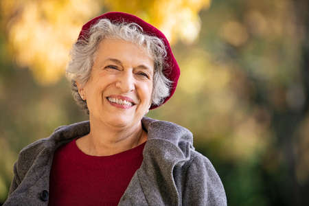 Portrait of senior woman smiling outdoor. Cheerful old woman in autumn park with copy space. Happy retired lady with grey hair and red beret smiling and looking away.の写真素材