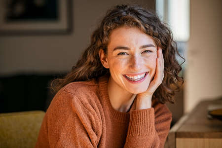 Portrait of beautiful young woman staying at home. Happy woman smiling while looking at camera with hand on cheek. Pretty laughing girl with curly hair and winter clothes relaxing in living room.の写真素材