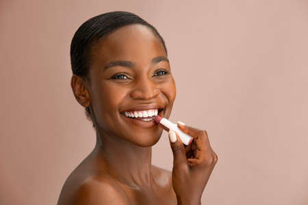 Cheerful beautiful woman applying natural lip balm brown background with copy space. Portrait of black woman applying brown lipstick while smiling. African american lady applying balm to her perfect lips isolated.の写真素材