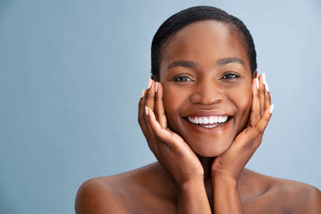 Portrait of beautiful african american woman touching her face and looking at camera. Close up face of black middle aged woman with bare shoulder massaging face isolated over blue background with copy space. Happy black lady touching soft cheek with clean and fresh skin.の写真素材