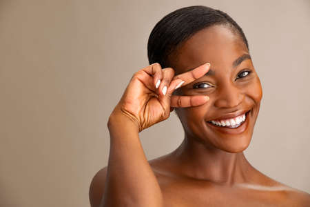 Portrait of smiling mature woman with perfect skin showing victory sign near eye on brown background. Closeup face of african woman with bare shoulder showing successful results after anti-aging wrinkle treatment. Crow's feet, eyebag and dark circles under the eyes concept.の写真素材