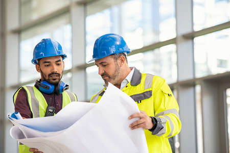 Young indian architect and mature supervisor meeting at construction site. Multiethnic manual worker and engineer wearing safety equipment and discussing on plan. Two construction workers working together while visiting a new building.の写真素材