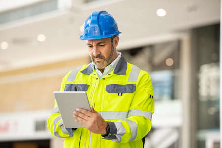 Serious and focused architect or engineer working on construction site using digital tablet while wearing safety vest and helmet. Mature man working on digital tablet at construction site. Mid adult manual worker with blue hardhat inspecting construction site while checking list on laptop.の写真素材