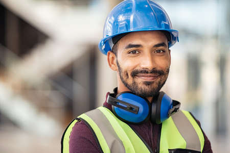 Portrait of satisfied construction site manager wearing safety vest and blue helmet at construction site. Young middle eastern architect watching construction site with confidence looking at camera. Indian manual worker on a industrial building with copy space.の写真素材
