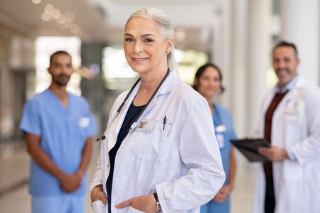 Portrait of mature female doctor on hospital corridor with hands in labcoat pockets and stethoscope. Confident general practitioner standing in hospital hallway with her healthcare team in background. Successful head physician in private clinic smiling and looking at camera.の写真素材