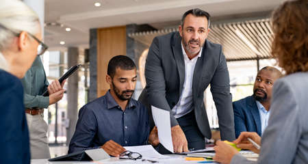 Multiethnic group of focused colleagues talking together during a brainstorming session. Group of businessmen and businesswomen listening to boss in meeting while discussion on new marketing strategy. Successful executive putting his ideas during presentation in conference room.の写真素材