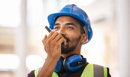 Young middle eastern bricklayer using walkie-talkie at construction site. Smiling indian site manager wearing helmet and yellow vest with safety ear muff while talking through walkie-talkie. Confident supervisor using walkie-talkie, giving instructions at construction site.の写真素材