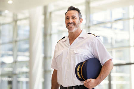 Smiling pilot in uniform standing at airport. Mature man wearing airplane uniform ready to fly at gate. Experienced commercial airplane pilot in uniform at modern airport smiling with copy space.の写真素材