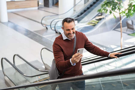 Smiling mid adult business man smiling while reading message on smartphone at airport. Successful senior casual man going up the escalator using smart phone while going to work. Businessman doing video call while standing in commercial building and using wireless earbuds.の写真素材