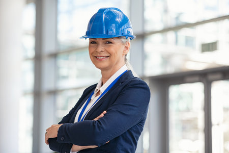 Portrait of smiling senior contractor wearing safety blue helmet looking at camera. Mature construction manager standing with crossed arms at site with copy space. Successful mid adult woman standing at building site while looking at camera with pride and satisfaction.の写真素材