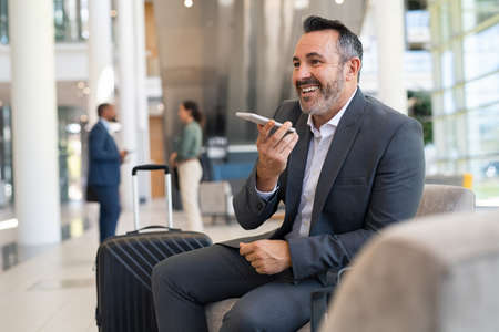 Smiling mature businessman sitting at airport lounge recording voice over smartphone for his colleague. Successful entrepreneur waiting at airport sending voice note. Businessman recording message while waiting his connection for business travel.の写真素材