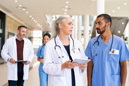 Mature woman physician discussing about medical report with young indian man nurse. Middle eastern nurse consulting senior doctor with patient details in hospital using digital tablet. Multiethnic team of general practitioners working together at medical clinic with copy space.の写真素材