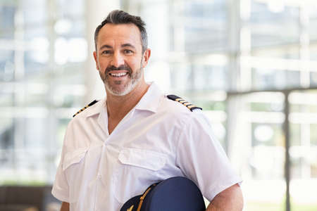 Smiling pilot in uniform standing at airport while looking at camera. Portrait of mature man wearing airplane uniform ready to fly. Successful mid adult pilot holding cap at gate with copy space.の写真素材