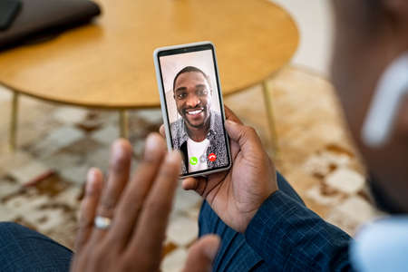 Close up of young african man on video call with boss using smartphone. Rear view of successful african business man taking interviewing a candidate over video call using mobile phone. Happy smiling client talking to financial advisor in a conference call.の写真素材