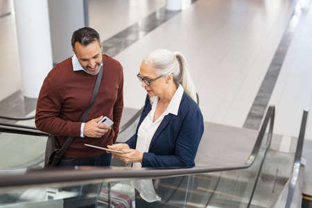 Businessman and woman talking on escalator using digital tablet while travelling. Mature businesswoman showing document on digital tablet to business man while moving on escalator at airport. Successful man listening to business woman while going to work.の写真素材
