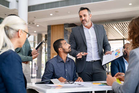Successful entrepreneur focusing on work with charts during meeting with multiethnic employee. Mature businessman discussing work at conference table in office with senior businesswoman. Office colleagues having discussion during meeting in conference room.の写真素材