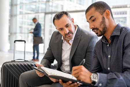 Two multiethnic businesspeople sitting at departure lounge at the airport discussing work while making notes in agenda. Indian business man working at airport with worried manager while waiting in lounge their connection. Successful businessman discussing during a business travel.の写真素材