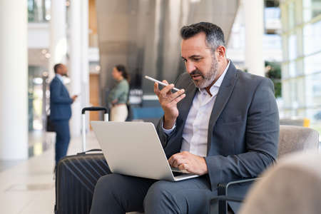 Serious businessman sitting on couch in airport lounge using laptop and sending voice message with smartphone. Mature business man working on computer while using voice command recorder on mobile phone. Mature businessman worried while talking at the phone.の写真素材