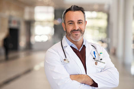 Portrait of happy doctor in labcoat and stethoscope standing with folded hands in modern hospital corridor. Successful general practitioner looking at camera with satisfaction at private clinic. Confident proud head physician standing in hospital hallway smiling with copy space.の写真素材