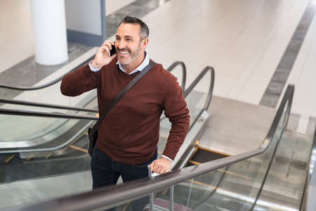 Smiling mature man standing in escalator while talking over smartphone at airport. Successful business man in airport standing on escalator while doing a conversation on the phone. Happy mid adult man with suitcase talking over mobile phone with colleague.の写真素材