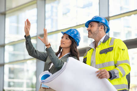 Young woman architect explaining blueprint to supervisor wearing safety vest at construction site. Mid adult contractor holding blueprint and understanding manager vision at construction site. Smiling engineer with hardhat on head talking to contractor while standing in building in construction process.の写真素材