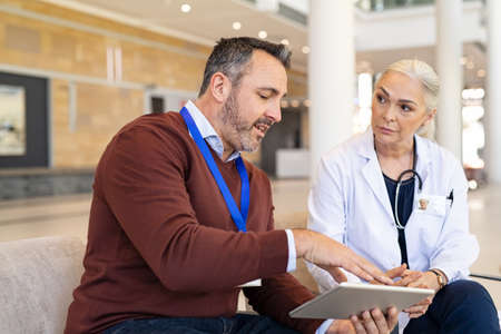 Pharmaceutical sales representative and mature woman physician having a discussion in hospital hallway. General practitioner discussing patient case status with his medical staff using digital tablet. Pharmaceutical representative showing medical report on digital tablet to mature doctor.の写真素材
