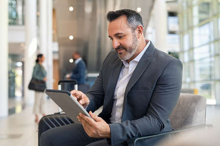 Successful business man sitting in airport lounge working on digital tablet. Mature businessman using digital tablet while sitting and waiting for boarding at international airport gate. Mid adult businessman in formalwear sitting in lounge of modern airport and working.の写真素材