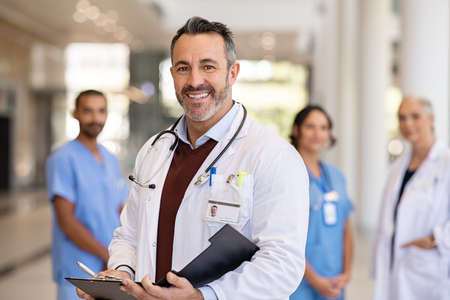 Portrait of handsome mature doctor wearing labcoat and stethoscope reading patient case file in hospital corridor. Successful head physician standing in hospital hallway and looking at camera with his medical team in background. Reliable general practitioner in career with his healthcare staff.の写真素材