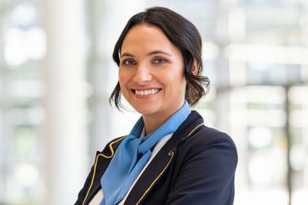 Portrait of beautiful stewardess standing at airport terminal waiting passengers. Charming flight attendant looking at camera, aircrafts are waiting and preparing flight. Portrait of smiling young woman flight attendant in dark blue uniform.の写真素材