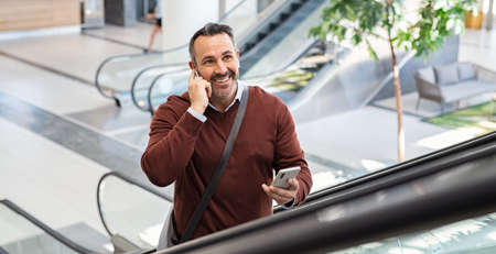 Smiling successful business man going up in escalator talking on smart phone while using wireless earbuds at airport. Casual mid adult man in modern building using cellphone while talking with colleague. Happy entrepreneur talking over mobile phone while going to work.の写真素材
