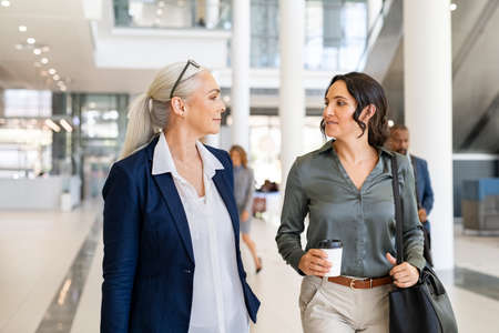 Two business women walking and talking in modern office. Two successful women manager discussing work while entering office building. Successful mature business woman talking with her assistant while going to work.の写真素材