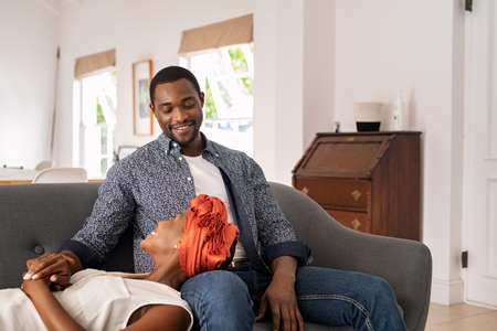 Mature african american woman lying on man's lap while talking with him on couch at home. Happy mid adult black wife lying on sofa while relaxing with husband. Smiling middle aged couple in conversation at home while looking at each other.の写真素材
