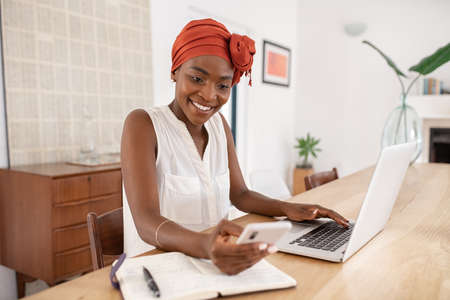 Smiling mature black woman using smartphone while working on laptop from home. African middle aged businesswoman checking notification on smart phone while taking a break during work. Successful black lady messaging with mobile phone while working at home: Smart working and telecommuting concept.の写真素材