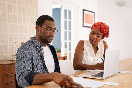Serious mid adult man and african woman with traditional turban working and writing notes while using laptop. Mature african american couple discussing home finance with bank statement and laptop. Black man and serious woman managing budget and checking accountancy and bills.の写真素材