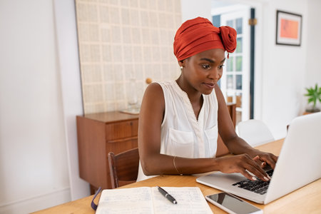 Mature black woman with traditional turban sitting at home while working on laptop. Successful black businesswoman typing on laptop wearing a traditional turban while working from home with copy space. Mid adult entrepreneur using computer during lockdown.の写真素材