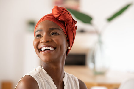 Beautiful mature black woman with african turban relaxing at home. Cheerful middle aged black woman in casual clothing with traditional headscarf at home laughing. African american lady smiling and looking away with copy space.の写真素材