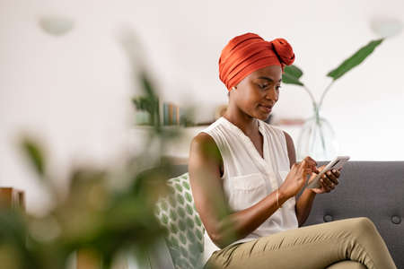 Mature african american woman sitting at home on couch using smart phone. Beautiful mid adult black woman typing on mobile phone. Smiling ethnic woman with traditional head turban using cellphone while sitting on sofa with copy space.の写真素材