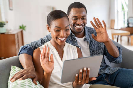 Smiling mature couple waving hands during video call using digital tablet. Middle aged black man and beautiful woman sitting on couch together doing video conference with family or friends on laptop. Cheerful mid adult husband and african american wife in conversation making video call with tablet.の写真素材