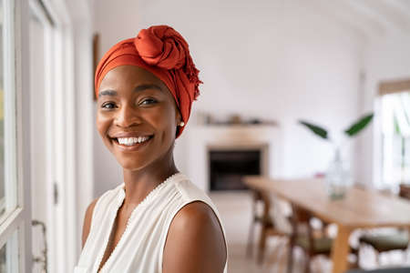 Portrait of a beautiful african woman smiling while looking at camera. Mid adult black woman in casual with traditional african headscarf stay at home and smiling. Cheerful mature lady standing near the window with copy space.の写真素材