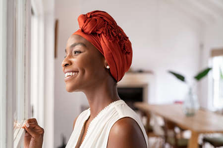 Smiling mid adult african american woman with turban looking outside the window. Cheerful black mature woman wearing traditional red headscarf while contempliting outdoor. Happy middle aged lady looking outside the window at home while thinking.の写真素材