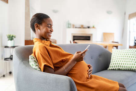 Mature black pregnant woman texting on mobile phone while sitting on couch in living room. Happy mid adult african american pregnant woman using smartphone at home. Beautiful lady messaging on cellphone while relaxing on sofa at home: motherhood and pregnancy concept.の写真素材