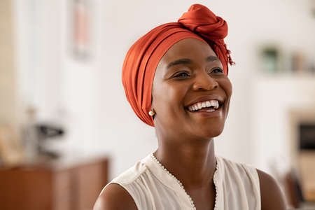Smiling middle aged African American woman with orange headscarf at home. Beautiful black woman in casual clothing with traditional turban at home laughing. Portrait of mature carefree lady smiling and looking away.の写真素材